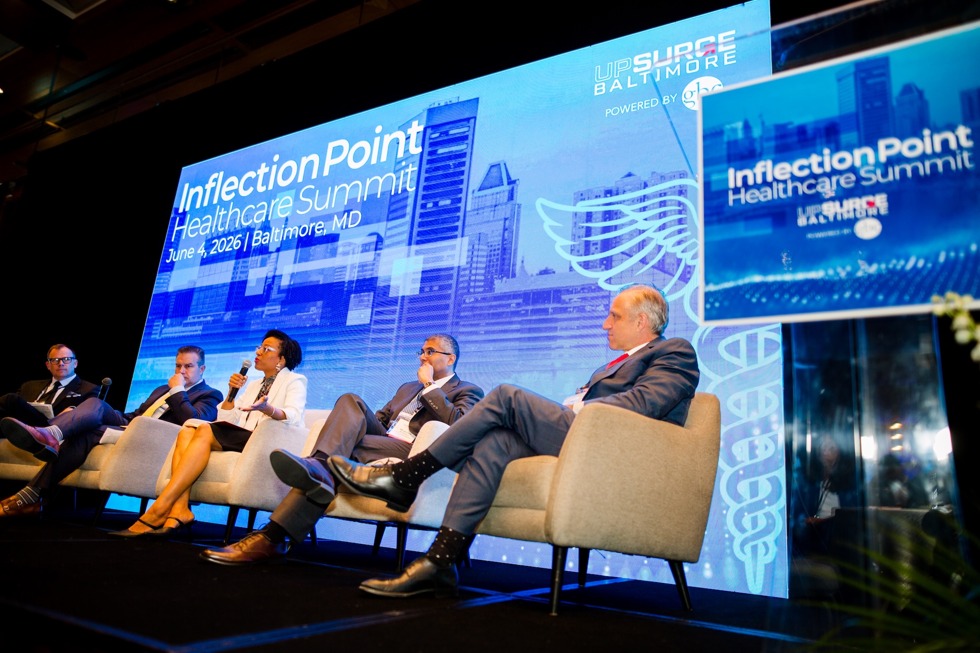 Five individuals seated in beige chairs with one in the center speaking as blue screens with white text and a city skyline superimposed within it broadcast behind and next to them with the words 